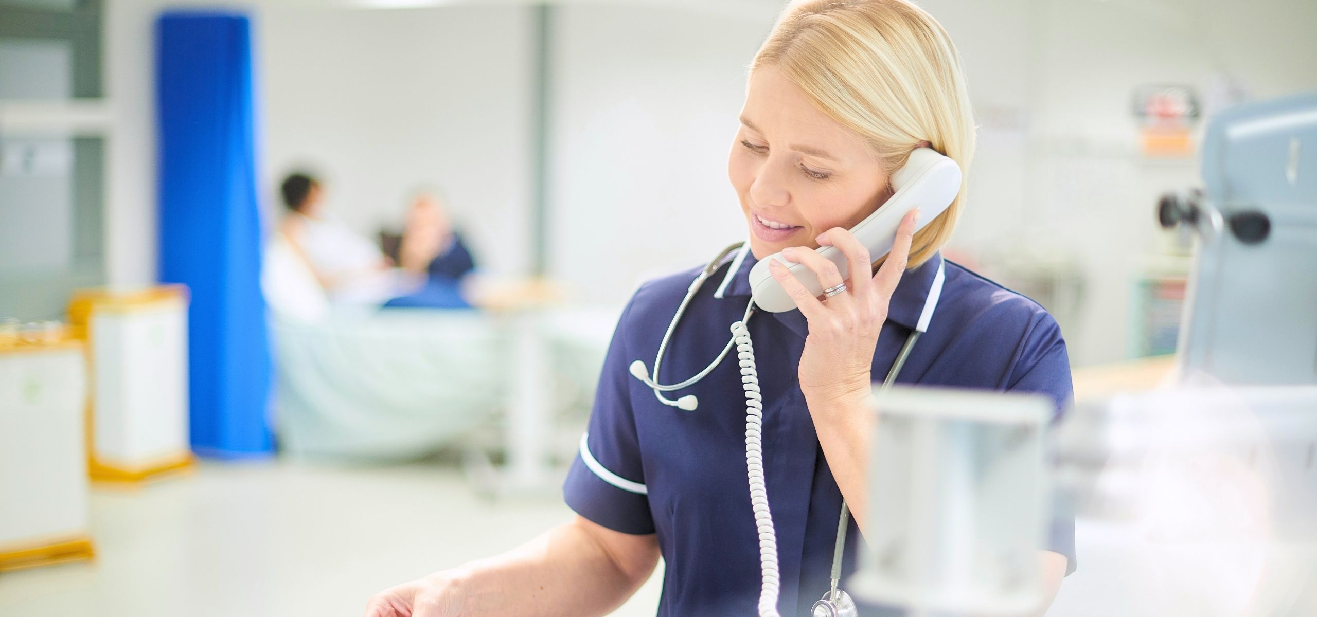 Nurse in scrubs with a stethoscope taking a phone call in a bright clinic hallway