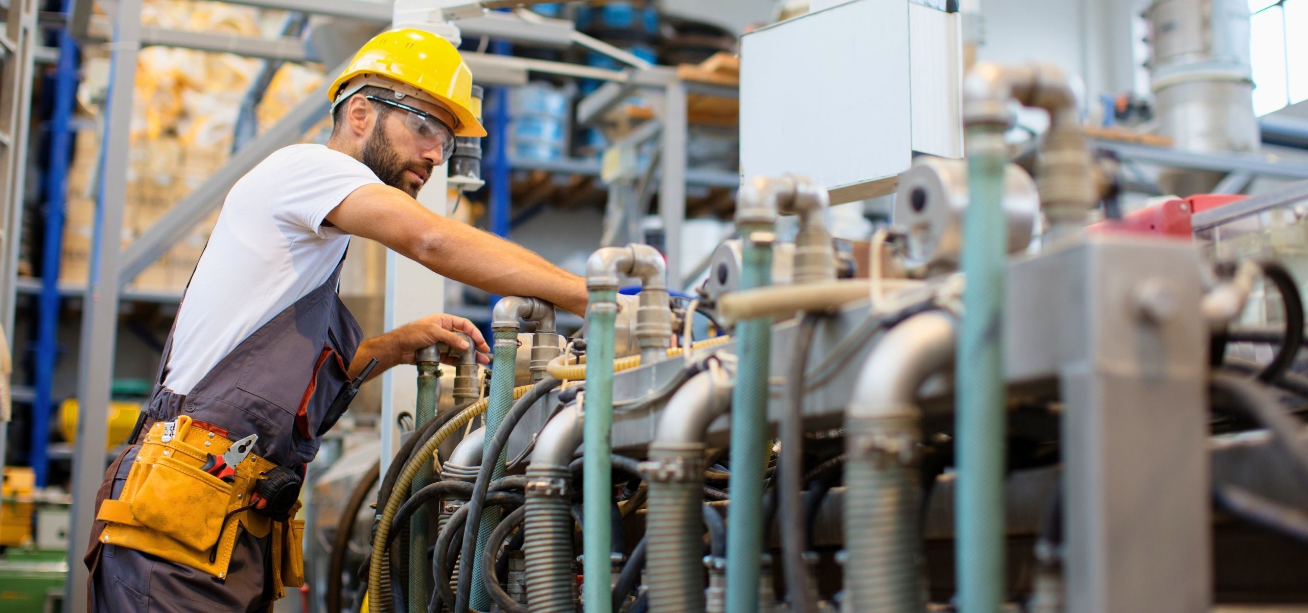 Industrial maintenance technician in a hard hat and tool belt servicing pipes and machinery in a plant