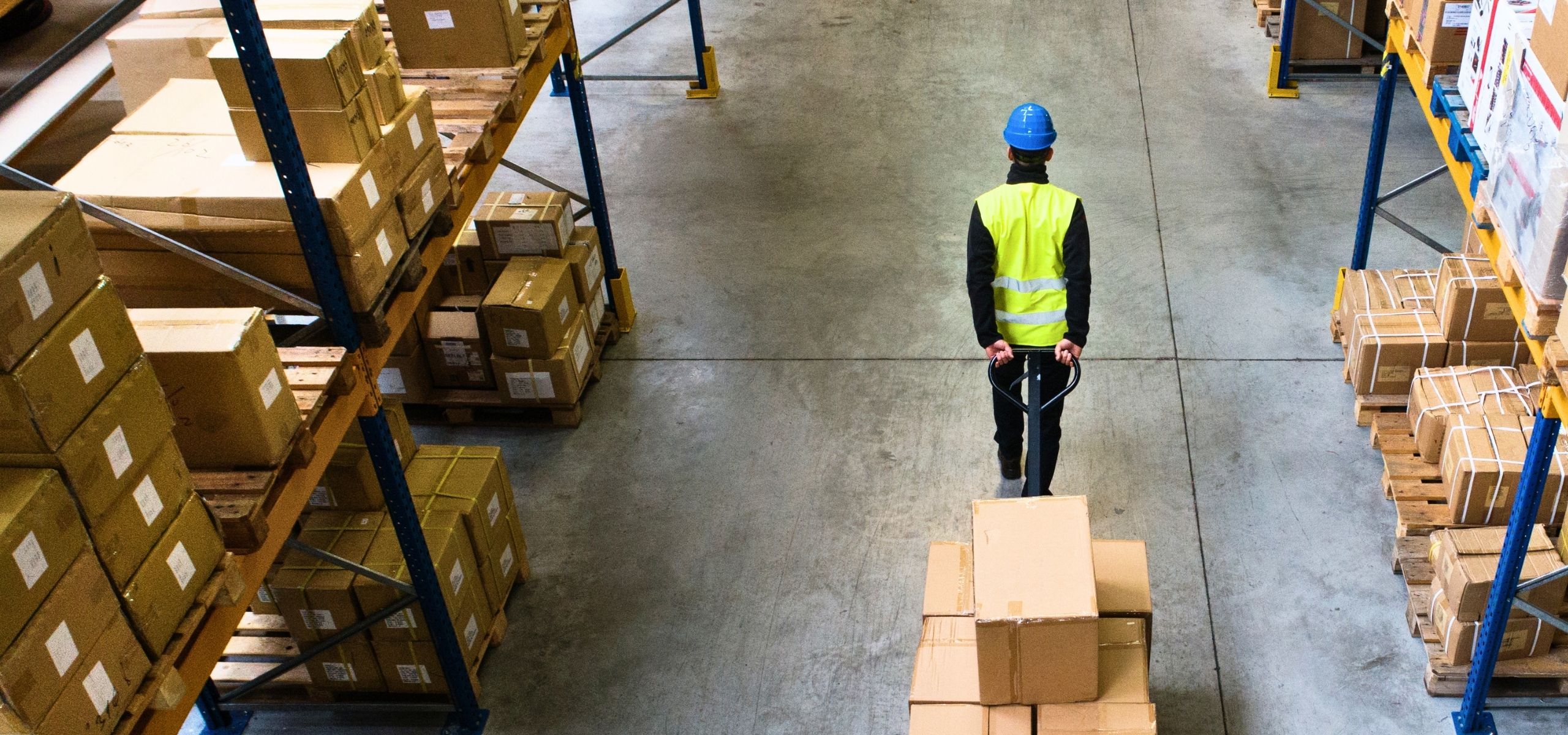 Warehouse associate in a safety vest moving a loaded pallet down a distribution center aisle